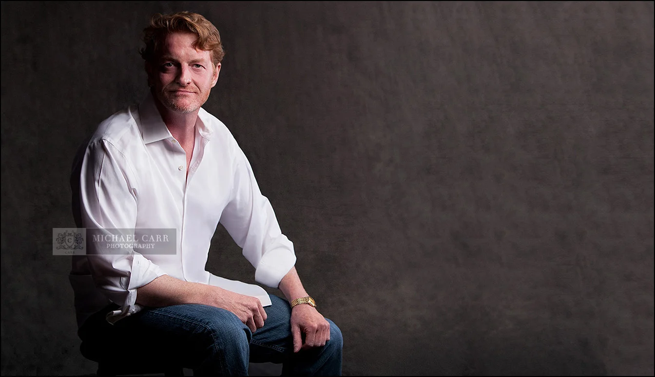 Personal branding portrait of a creative professional seated against a textured dark backdrop.