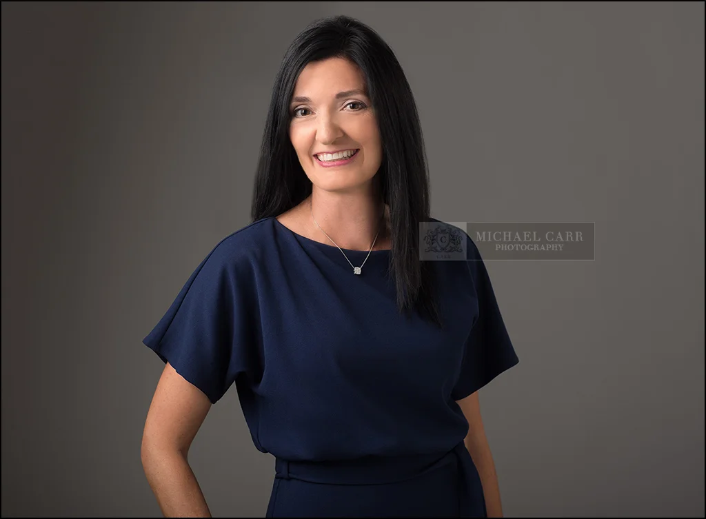 Houston Headshots Confident businesswoman in a navy dress smiling against a gray studio background.